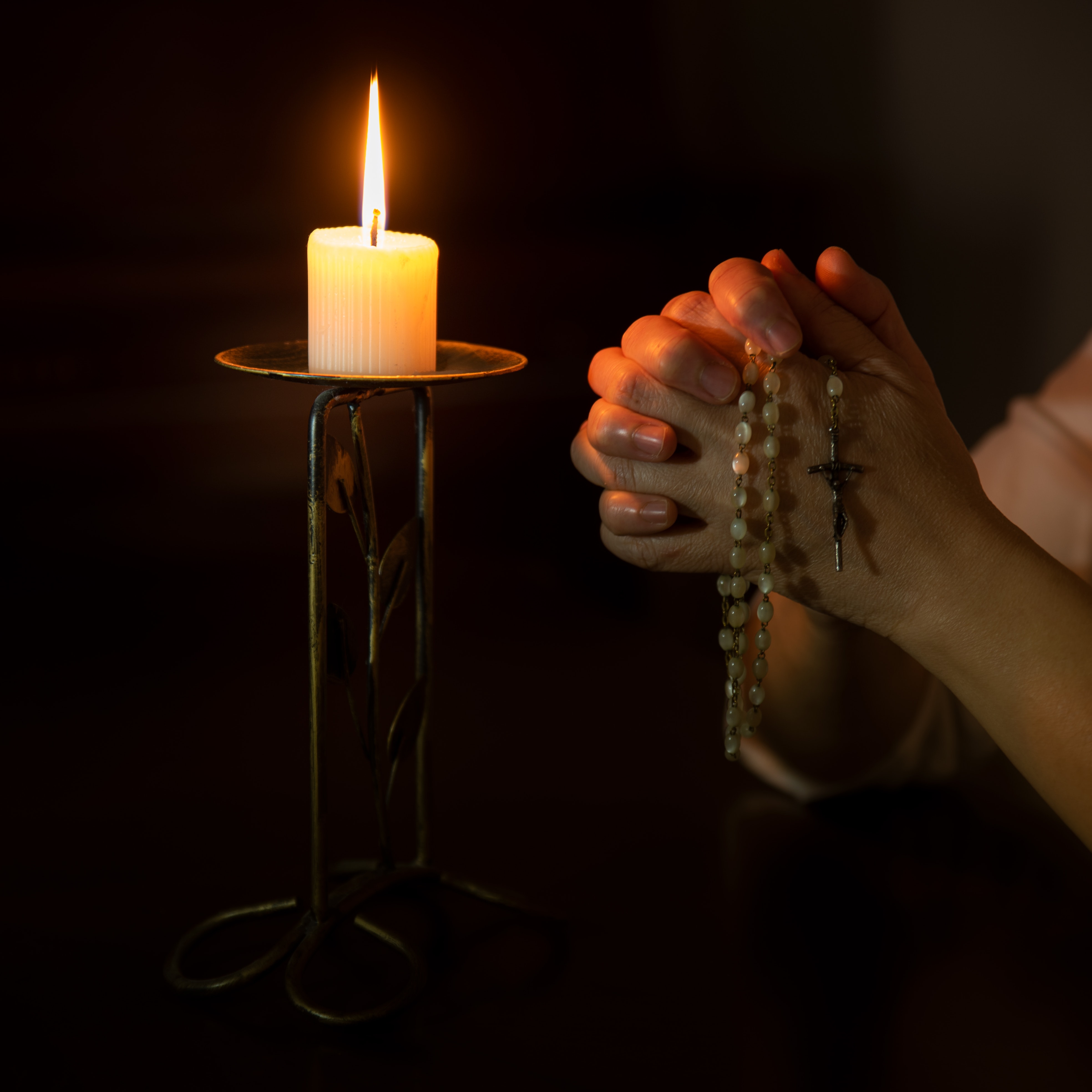 Praying hands holding a rosary, next to a lit candle